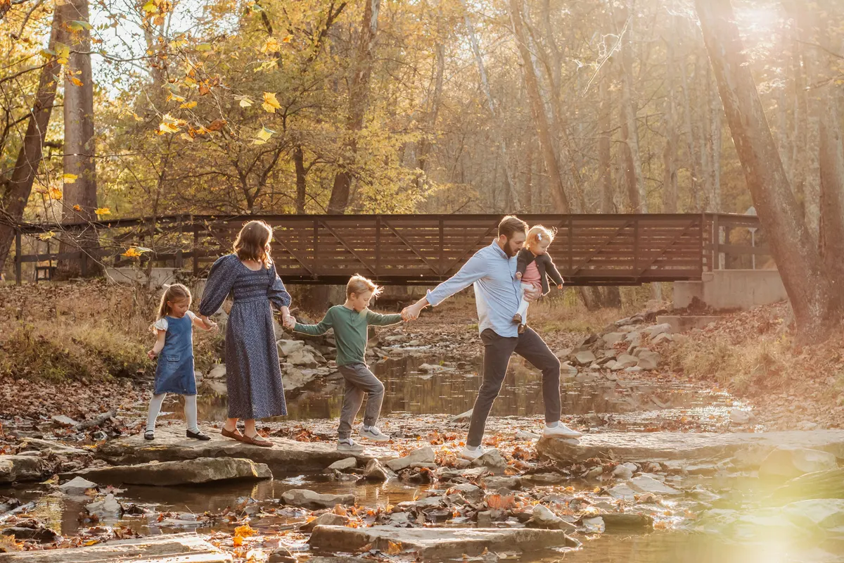 Family crossing creek stones together near a wooden footbridge in fall